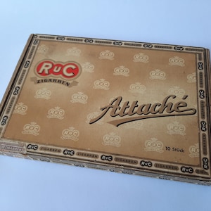 May include: A vintage box of RUC Zigarren cigars. The box is tan with a repeating pattern of the RUC logo and the word "Attaché" in a cursive font. The box also has the text "10 Stück".