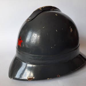K&ouml;nnte beinhalten: Ein dunkelgrauer Vintage-Helm mit einem markanten Mittelkamm und einem kleinen roten Stern-Emblem an der Seite. Der Helm hat eine gl&auml;nzende Oberfl&auml;che mit sichtbaren Gebrauchsspuren, einschlie&szlig;lich Farbabplatzungen an den Kanten.