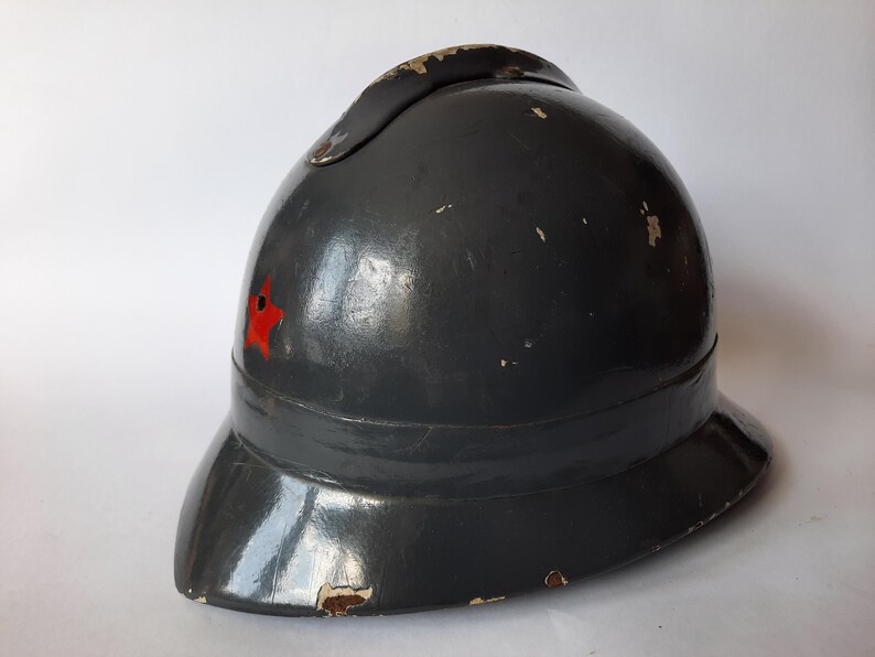 K&ouml;nnte beinhalten: Ein dunkelgrauer Vintage-Helm mit einem markanten Mittelkamm und einem kleinen roten Stern-Emblem an der Seite. Der Helm hat eine gl&auml;nzende Oberfl&auml;che mit sichtbaren Gebrauchsspuren, einschlie&szlig;lich Farbabplatzungen an den Kanten.