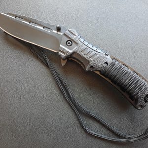Peut inclure: Un couteau pliant noir avec une poignée texturée et un cordon noir. La lame est dentelée et possède une soie partielle.