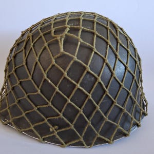 Puede incluir: Un casco de estilo militar de color verde oliva oscuro con forma de cúpula redondeada y ala ancha. El exterior del casco está cubierto por una red de cuerda gruesa de color beige con un patrón de rombos que se extiende por toda la superficie.