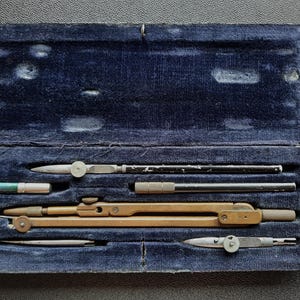 Pode incluir: Um estojo vintage de veludo azul aberto, contendo várias ferramentas de desenho. As ferramentas incluem um grande compasso de latão, um pequeno cilindro verde e prateado e outros instrumentos de metal. O estojo está sobre uma superfície escura.
