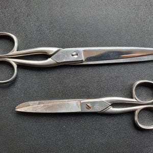 Puede incluir: Dos pares de tijeras plateadas vintage con orificios redondeados para los dedos. Las hojas son afiladas y las tijeras están sobre una superficie negra texturizada. Un par es más grande que el otro.