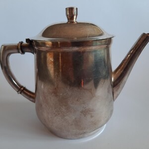 May include: A silver-colored teapot with a lid and a curved spout. The teapot has a handle and a small knob on the lid. The surface of the teapot has a slightly aged appearance, suggesting it is vintage or antique. The teapot is likely used for serving tea.