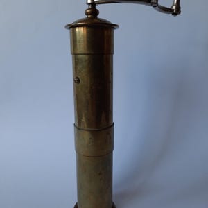 Puede incluir: Un molinillo de pimienta vintage de latón con una manivela de plata. El molinillo tiene una forma cilíndrica y un acabado liso y envejecido.