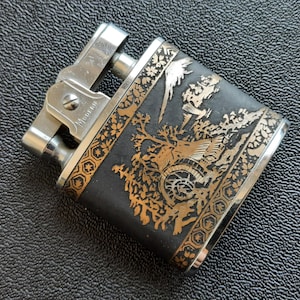 May include: Vintage lighter with a silver metal body and a black and gold engraved design. The design features a detailed landscape scene. The word "Modern" is engraved on the lighter.