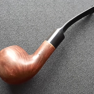 Pode incluir: Um cachimbo de tabaco curvo com uma tigela castanha e um bocal preto. O cachimbo tem uma faixa de madeira e o texto "BARASSO" é visível na lateral da tigela. O cachimbo está sobre uma superfície cinza escuro texturizada.