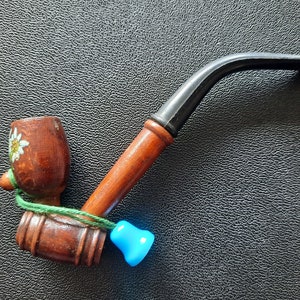 May include: A wooden tobacco pipe with a dark brown bowl and stem, featuring a light brown handle. The bowl is decorated with a painted flower. A blue bell-shaped charm is attached with green string.