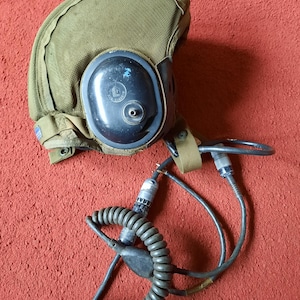 Può includere: Un casco da aviatore verde oliva con auricolare nero e cavo di comunicazione a spirale. Il casco ha una copertura in tessuto e un cinturino. L'auricolare ha un design circolare con un piccolo foro.