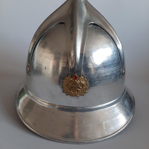 Può includere: Un casco da pompiere vintage color argento con una parte superiore arrotondata e un'ampia tesa. Il casco presenta un emblema dorato decorativo con un accento rosso. Il casco è in metallo e ha una superficie riflettente.