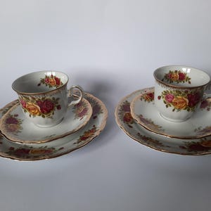 May include: Two sets of vintage teacups, saucers, and side plates. Each set features a white background with a floral rose pattern in shades of red, yellow, and pink. The edges are trimmed with gold. A classic tea set.