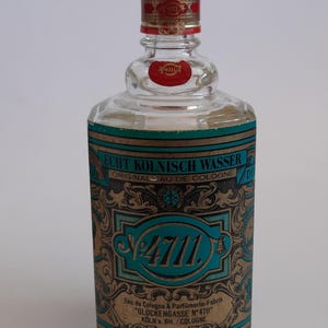 Puede incluir: Una botella de vidrio transparente de "4711" Eau de Cologne. La botella tiene una tapa dorada con una banda roja y una etiqueta roja. La etiqueta de la botella es verde azulado y dorada con el texto "Echt K&ouml;lnisch Wasser" y "Original Eau de Cologne".