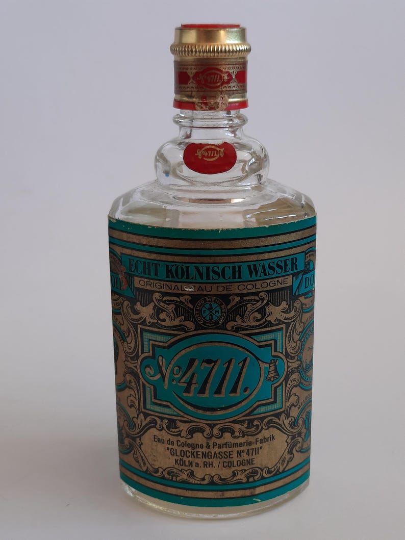 Puede incluir: Una botella de vidrio transparente de "4711" Eau de Cologne. La botella tiene una tapa dorada con una banda roja y una etiqueta roja. La etiqueta de la botella es verde azulado y dorada con el texto "Echt K&ouml;lnisch Wasser" y "Original Eau de Cologne".