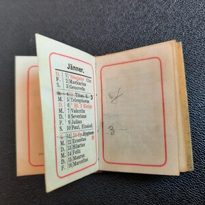 Puede incluir: Un peque&ntilde;o libro encuadernado en papel blanco con un borde rojo alrededor de los bordes. El libro est&aacute; abierto en una p&aacute;gina con un calendario para el mes de enero. El calendario est&aacute; en alem&aacute;n y enumera los d&iacute;as de la semana y las fechas. El texto de la p&aacute;gina incluye los nombres de los santos y las festividades.