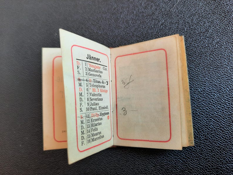 Puede incluir: Un peque&ntilde;o libro encuadernado en papel blanco con un borde rojo alrededor de los bordes. El libro est&aacute; abierto en una p&aacute;gina con un calendario para el mes de enero. El calendario est&aacute; en alem&aacute;n y enumera los d&iacute;as de la semana y las fechas. El texto de la p&aacute;gina incluye los nombres de los santos y las festividades.