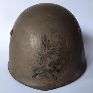 Può includere: Un elmetto color verde oliva, usurato, con una corona arrotondata. L'elmetto presenta due rivetti laterali e un disegno sbiadito di impronta di mano nera sul davanti. La superficie mostra segni di usura.