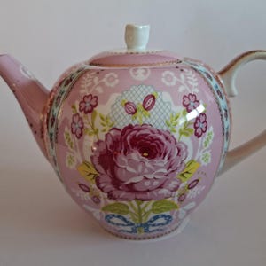 Puede incluir: Una tetera de cerámica rosa con una gran ilustración central de una rosa de color rosa oscuro, hojas verdes y un lazo azul en la base. La tetera tiene una tapa a juego con un pomo blanco, un asa curva y un pico, todo con detalles en dorado.