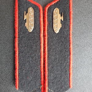Könnte beinhalten: Ein Paar schwarze Schulterklappen mit roter Paspel und einem goldenen Panzerabzeichen. Das Panzerabzeichen ist eine stilisierte Darstellung eines Panzers mit einem Turm und Rädern.