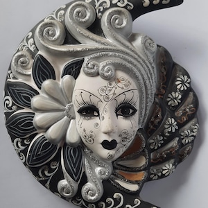 Peut inclure: Un masque mural décoratif présentant un visage central avec des détails peints en noir et blanc, entouré d'une forme de croissant de lune. Le design comprend des accents de paillettes argentées, des motifs floraux et des volutes en noir et blanc.