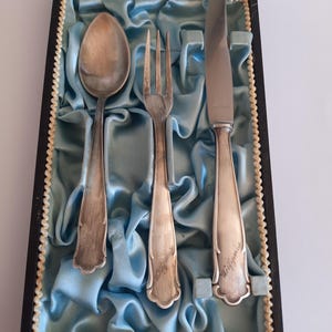 May include: A vintage silverware set in a black box with a light blue satin lining. The set includes a spoon, fork, and knife. The silverware is silver-colored, and the knife has the name "Wolfgang" engraved on it.