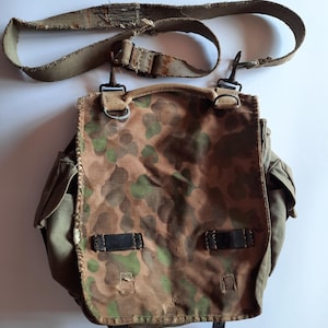 Puede incluir: Un bolso mensajero de camuflaje vintage con una correa de lona desgastada y herrajes metálicos. El bolso presenta un cierre de solapa con detalles de cuero negro y bolsillos laterales. El patrón de camuflaje incluye tonos de marrón, verde y beige.