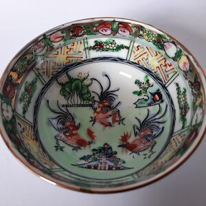 May include: A small, round, antique porcelain bowl with a gold rim. The interior features a light green background with three red and black rooster illustrations. The rim is decorated with floral and geometric patterns in various colors.