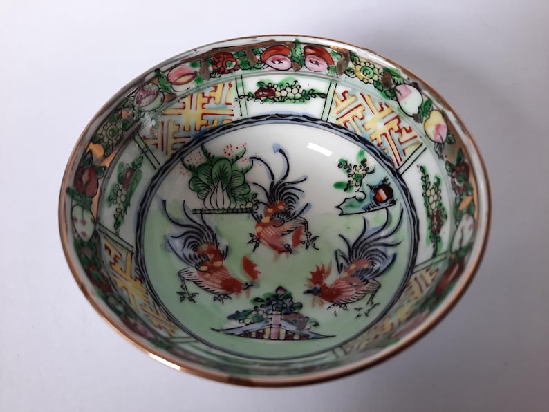 May include: A small, round, antique porcelain bowl with a gold rim. The interior features a light green background with three red and black rooster illustrations. The rim is decorated with floral and geometric patterns in various colors.