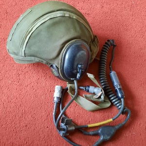 Könnte beinhalten: Ein olivgrüner Militärhelm mit einem schwarzen Headset und einem spiralförmigen Kommunikationskabel. Der Helm hat eine gepolsterte Innenseite und eine Stoffaußenseite. Das Headset hat ein Mikrofon und einen Ohrmuschel. Das Kabel hat mehrere Anschlüsse.