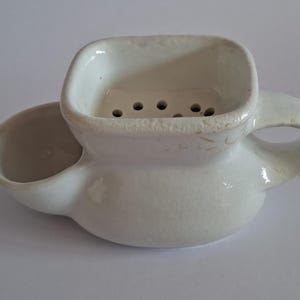 Może przedstawiać: Biały ceramiczny kubek do golenia z zaokrąglonym bocznym zbiornikiem oraz kwadratową górną komorą z perforowaną podstawą zawierającą kilka małych otworów odpływowych. Naczynie posiada zakrzywiony uchwyt umieszczony po prawej stronie.