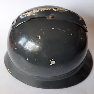 K&ouml;nnte beinhalten: Ein dunkelgrauer, kuppelf&ouml;rmiger Metallhelm mit einer breiten, ausgestellten Krempe. Die Oberfl&auml;che weist ein gl&auml;nzendes Finish mit sichtbaren Lackabplatzungen und Kratzern auf, insbesondere am oberen Grat, wo ein Metallstreifen vernietet ist.