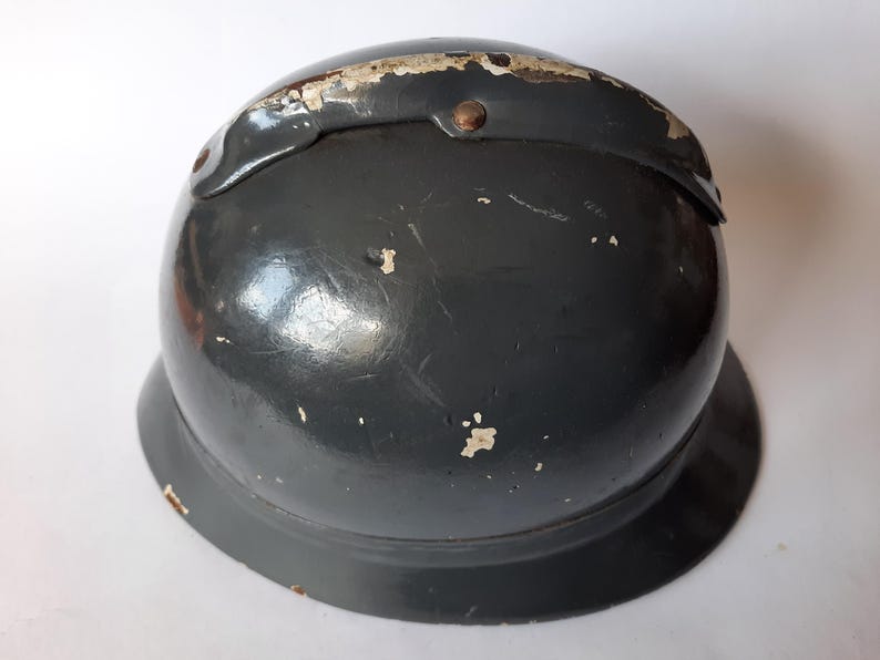K&ouml;nnte beinhalten: Ein dunkelgrauer, kuppelf&ouml;rmiger Metallhelm mit einer breiten, ausgestellten Krempe. Die Oberfl&auml;che weist ein gl&auml;nzendes Finish mit sichtbaren Lackabplatzungen und Kratzern auf, insbesondere am oberen Grat, wo ein Metallstreifen vernietet ist.