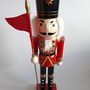 May include: A wooden nutcracker figure wearing a black hat with a gold star and red gem, a red jacket with green rhinestones, and black boots. The figure has white hair and a beard, and holds a tall pole with a red fabric flag in its right hand.