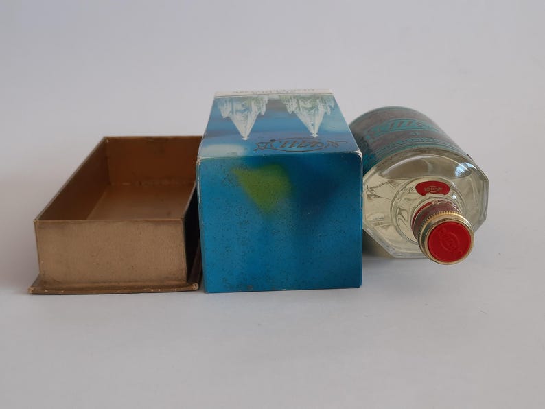 Puede incluir: Una botella de vidrio transparente con tapa roja y etiqueta azul y blanca, junto a una caja azul con dise&ntilde;o de monta&ntilde;a y una peque&ntilde;a caja marr&oacute;n abierta. La botella est&aacute; tumbada de lado.