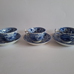 May include: Three white ceramic teacups and matching saucers featuring a blue transferware pattern. The design depicts a landscape with a horse-drawn carriage, trees, and floral motifs. Each cup has a curved handle and a flared rim.
