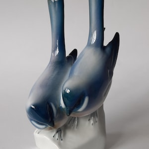 Può includere: Una statuetta in porcellana di due uccelli blu appollaiati su una base bianca. Gli uccelli si guardano e hanno una finitura smaltata.