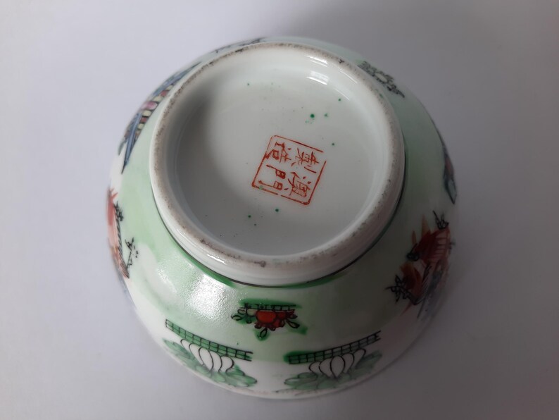 May include: An antique porcelain bowl with a light green exterior and a white interior. The bowl features colorful painted designs of flowers, birds, and other decorative elements. The bottom of the bowl has a red stamp.