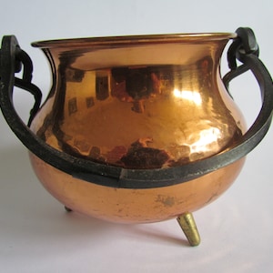 May include: A small copper pot with three brass feet and two black iron handles. The pot has a hammered finish and is slightly dented.
