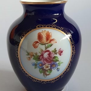 Peut inclure: Un vase en porcelaine bleu cobalt avec un médaillon circulaire blanc central orné d'un bouquet floral coloré composé de roses et de petites fleurs. Le vase présente un bord doré et une bordure en filigrane doré autour du motif floral.