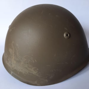 Può includere: Un elmetto militare verde oliva con la parte superiore arrotondata e un bordo leggermente curvo. L'elmetto presenta alcuni graffi e un piccolo rivetto laterale. L'elmetto è probabilmente in metallo ed è progettato per la protezione.