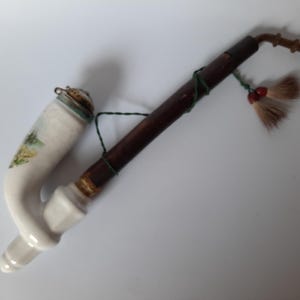 Op de afbeelding: Een vintage tabakspijp met een witte keramische kom met een landschapsontwerp. De steel is donkerbruin met een decoratieve kwast. Een klassiek verzamelobject.