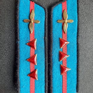 May include: Pair of vintage blue shoulder boards with red stripes and red triangle accents. Each board features a gold-colored emblem and black trim. A classic military or uniform accessory.
