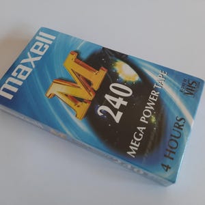 Könnte beinhalten: Ein blaues und goldenes Maxell VHS-Videokassettenband mit der Aufschrift "Mega Power Tape" und dem Text "240" und "4 Stunden" auf der Vorderseite. Das Band ist mit "E-220M" und "VHS" mit einem Logo beschriftet.