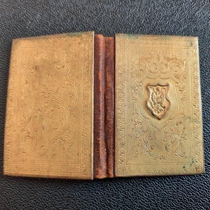 Puede incluir: Un peque&ntilde;o libro dorado con bisagras, con un dise&ntilde;o ornamentado y un escudo en la portada.