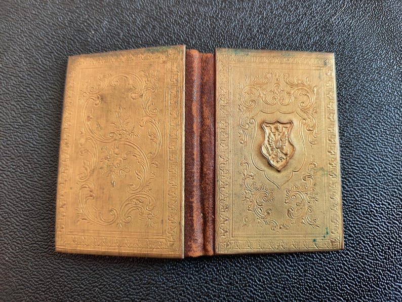 Puede incluir: Un peque&ntilde;o libro dorado con bisagras, con un dise&ntilde;o ornamentado y un escudo en la portada.
