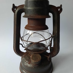 Könnte beinhalten: Eine antike, rostige, grüne Metall-Petroleumlampe mit einer Glaskugel und einem Holzgriff. Die Lampe hat einen Metallgriff und einen Metallkäfig um die Glaskugel. Die Lampe trägt den Text "No. 225" auf der Seite.