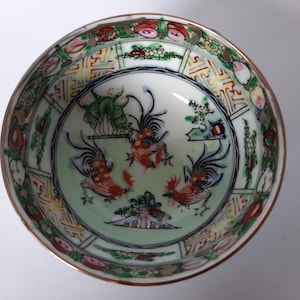 May include: A decorative porcelain bowl with a detailed hand-painted design. The bowl features a central image of three roosters in shades of red, orange, and blue, surrounded by floral and geometric patterns. The rim is trimmed with gold.