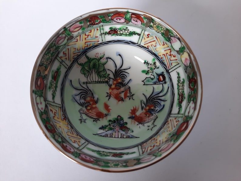 May include: A decorative porcelain bowl with a detailed hand-painted design. The bowl features a central image of three roosters in shades of red, orange, and blue, surrounded by floral and geometric patterns. The rim is trimmed with gold.