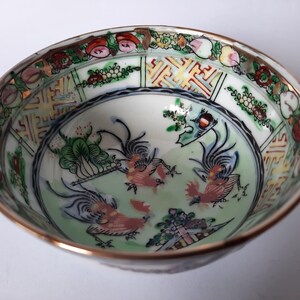 May include: A detailed, antique porcelain bowl with a gold rim. The interior features a light green background with three stylized rooster figures in red and white. The rim is decorated with floral and geometric patterns in various colors.