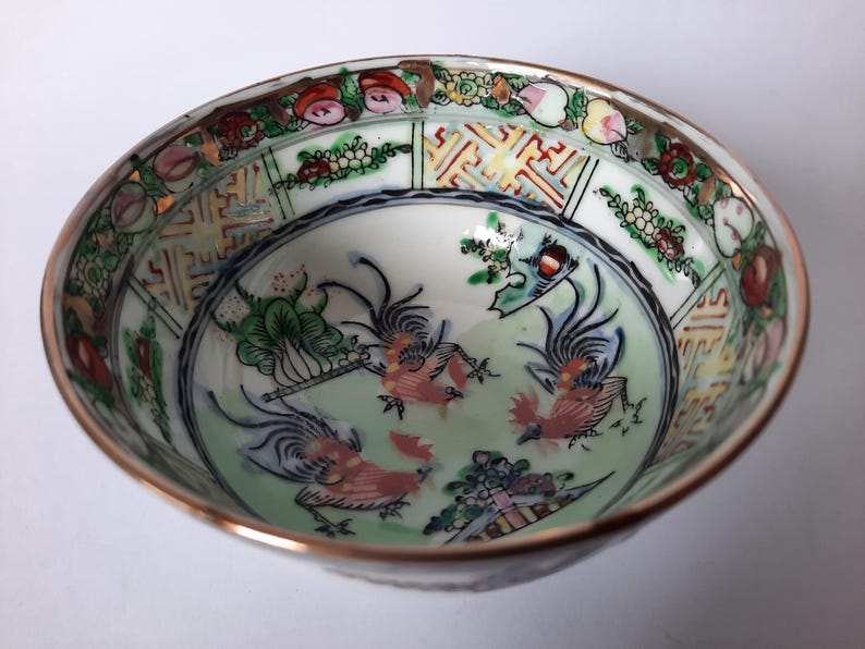 May include: A detailed, antique porcelain bowl with a gold rim. The interior features a light green background with three stylized rooster figures in red and white. The rim is decorated with floral and geometric patterns in various colors.