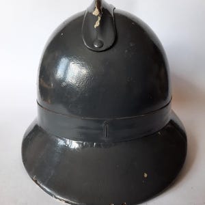K&ouml;nnte beinhalten: Ein dunkelgrauer, gl&auml;nzender, abgerundeter Helm mit einem zentralen vertikalen Grat an der Oberseite. Der Helm hat eine breite, ausgestellte Krempe und weist sichtbare Gebrauchsspuren auf, darunter Farbabsplitterungen am Kamm und am Rand.